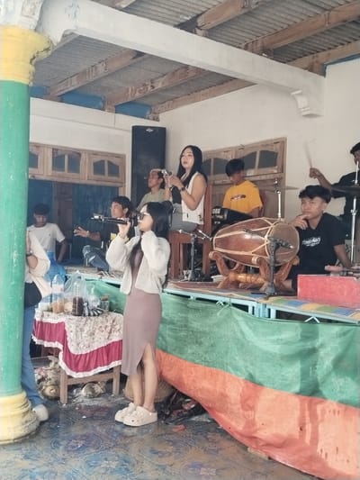 The image depicts a musical performance in what appears to be an outdoor or semi-outdoor setting in Jenawi, Indonesia. A woman wearing a light cardigan, a dress with a slit, and light-colored clogs is singing into a microphone. Behind her, a band is performing with various instruments including a traditional Indonesian drum (kendang), keyboard, and drums. Other people are visible, either as part of the band or observing the performance. There are drinks and water bottles on a table in front of her. The stage is covered in green and red material. The architectural style of the building or structure suggests a local Indonesian design.
