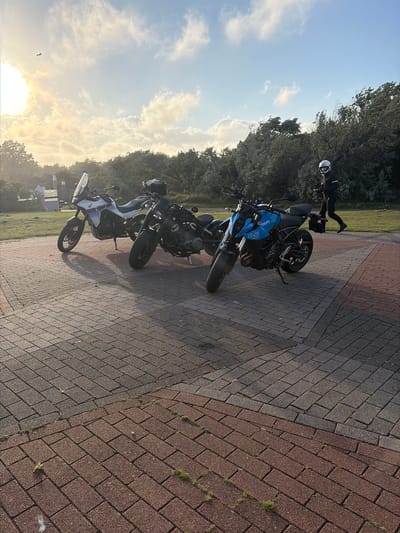 The image depicts an outdoor scene in the city of Hook of Holland, Netherlands, likely during the early morning or late afternoon due to the low angle of the sun casting long shadows. Two motorcycles are parked on a paved area made of red and grey interlocking bricks.

In the foreground, a blue Suzuki motorcycle with "8S" branding is prominently displayed. Behind it, a white and blue adventure-style motorcycle with a tall windshield is parked. To the right of the motorcycles, a person dressed in full motorcycle riding gear, including a white helmet and black jacket and pants, is walking away from the viewer towards the right of the frame. The person is holding what appears to be a mobile device or navigation unit in their left hand.

The background features a lush green grassy area with a line of dense trees and shrubs. In the far distance, the sky is visible with scattered clouds and the sun creating a bright halo effect. A bird is silhouetted against the bright sky in the upper left portion of the image. A light pole and a structure resembling an ice cream cone dispenser are faintly visible in the left mid-ground. The overall environment suggests a public area, possibly a viewpoint or a rest stop, given the presence of seating structures and the open expanse.