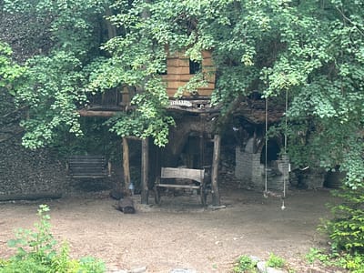 The image shows a rustic treehouse nestled amongst lush green foliage, located in the city of Golyamo Belovo, Bulgaria. The treehouse structure, made of rough-hewn wood and possibly straw-like material, has at least two square windows. It is supported by thick tree branches.
In front of the treehouse, a wooden swing set with a bench is positioned on the dirt ground. Another, simpler swing made of rope and a wooden seat hangs from a branch to the right, near a brick outdoor oven or fireplace structure. To the left, a suspended wooden bench swing is also visible, set against a backdrop of a rocky slope.
Several logs are scattered on the ground. A plastic bottle is placed near one of the support posts of the swing set. The ground is predominantly dirt and sand, with some sparse green vegetation growing in the foreground and around the edges of the scene. The overall impression is one of a natural, somewhat wild, and secluded recreational area.
