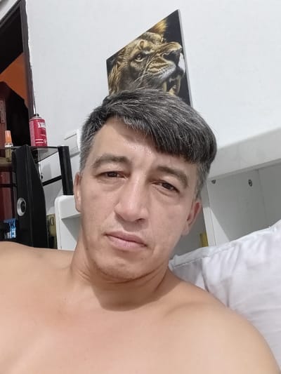 A close-up, eye-level selfie shows a fair-skinned man in his late 40s to early 50s with short, dark graying hair and faint stubble on his chin. He has light brown eyes and is looking directly at the camera with a neutral expression. His bare shoulders and upper chest are visible.
In the background, a white wall is adorned with a framed picture of a lion's head. To the left, a dark-colored shelf holds a red aerosol can and a glass bottle. A portion of a doorway with an orange interior is also visible. A white pillow is prominent on the right side of the frame. The scene appears to be indoors, possibly a bedroom. The location is not specified within the image.