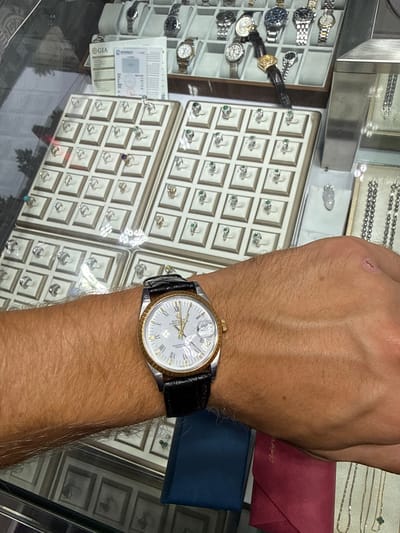 A person's wrist wears a gold-toned watch with a black leather band, positioned in front of a display case filled with jewelry. The watch face is white with Roman numerals and appears to be a Rolex Oyster Perpetual. Behind the watch, several trays hold rings, some with green gemstones. In the background, other jewelry, including watches, necklaces, and pendants, are displayed. A GIA (Gemological Institute of America) certificate is visible on a stand. The scene is indoors, likely a jewelry store in Liwan District, China.