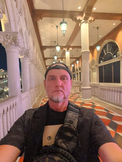 The media file shows a selfie of a man in what appears to be an indoor setting with architectural details reminiscent of old European or Venetian style. The man is wearing a black t-shirt with a camouflage vest over it and a black cap. He has a goatee and is looking towards the camera. The background features ornate columns and railings, possibly from a balcony or walkway. The floor has a distinctive diamond pattern with orange, grey, and white tiles. Hanging lamps with green glass shades are visible, suggesting an interior space with decorative lighting. The overall impression is that of a tourist taking a photo at a hotel or resort, possibly in the evening or at night given the interior lighting and the distant view which seems to be illuminated buildings against a dark sky. The man's expression is neutral. There is text on a light brown tag visible on the man's shirt, but it is not clearly legible. The location is identified as Paradise, United States of America.