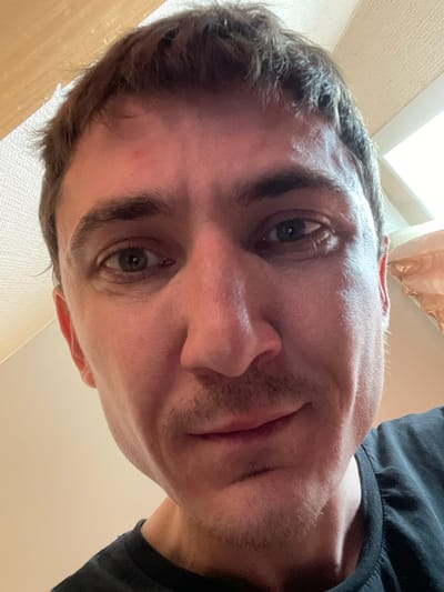 The image is a close-up, low-angle, Dutch-angle photograph of a person's face, with their head tilted downwards. The person appears to be a male with short, dark hair and light skin. They have a light stubble on their face. The visible eye is a greenish-brown color. The person is wearing a black t-shirt.
The background suggests an indoor setting, possibly a room, with a textured wall and a light-colored ceiling or upper wall visible. There's also a hint of what could be a window or a light source in the upper left corner, casting bright light. The overall lighting suggests it could be daytime, but the indoor setting makes this uncertain.
The photograph seems to be taken from a very close perspective, looking up at the person's face. The tilted angle of the image and the downward gaze of the person could imply a casual or spontaneous moment, perhaps a selfie taken from an unusual angle. No specific activity is clearly depicted, other than the person being present and looking towards the camera. There is no visible text in the image. The location context of Laishevo, Russia, does not provide any visual cues within the image itself.