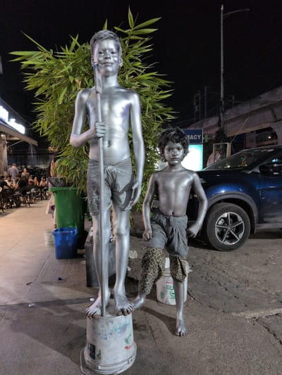 The scene depicts two silver-painted male children posing as living statues on a bustling urban street at night, consistent with the city of Hyderabad, India.

In the foreground, a taller male child stands to the left, entirely covered in silver paint, including his hair. He wears silver-painted shorts/loincloth and holds a long, thin, silver-painted staff vertically in front of him with both hands. His feet are bare, and he stands on an inverted, white, paint-splattered plastic bucket. He exhibits a slight smile and looks directly at the viewer. Behind him is a dense, tall clump of green foliage resembling bamboo.

To the right of the taller child is a shorter male child, also fully covered in silver paint, including his hair. He wears silver-painted shorts/loincloth and patterned, silver-painted leg coverings extending up his calves. He stands barefoot on the ground, with his right hand slightly forward and his left hand near his hip. He maintains a neutral expression, looking towards the viewer. Behind his left leg, a white plastic bucket with the "Amul Curd" logo is visible.

In the mid-ground, to the left, an outdoor dining area of a commercial establishment is visible under an awning, with several individuals seated at tables. A large green plastic dustbin stands between this area and the taller child. To the right, behind the shorter child, a dark blue SUV is parked, partially visible, along with a neon blue sign displaying "PHARMACY" with a white cross.

The background features a tall street light pole on the right side, and an elevated concrete structure, likely a metro rail or flyover, with utility wires, extends across the upper right. The ground transitions from a paved sidewalk on the left to a rougher asphalt or concrete surface on the right. A blue plastic bucket is also visible on the ground behind the taller child's legs.