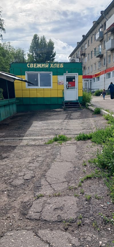 This media file captures an outdoor scene in Volsk, Russia. The primary subjects are a small, brightly colored kiosk, a residential building, and a paved area with some greenery.
The kiosk is painted in alternating horizontal stripes of green and yellow. It has a white door with a window and a sign above it. Below the door, there's a small window, and another larger window is visible on the side of the kiosk. Text in green lettering is visible on the side of the kiosk, which reads "СВЕЖИЙ ХЛЕБ" (Fresh Bread). Inside the kiosk, through the door's window, a red sign is partially visible.
Adjacent to the kiosk is a multi-story residential building. It appears to be constructed with brick and has numerous windows. The building is situated on a slight incline relative to the kiosk.
The ground in front of the kiosk is paved, showing signs of wear and tear with cracks and patches of gravel. Small green plants are growing through some of these cracks.
In the background, a large tree with dense green foliage is visible, and further back, more trees and the sky with clouds can be seen. The sky suggests it is daytime, likely overcast or partly cloudy.
Two individuals are partially visible in the upper left quadrant of the image, near the residential building. One is wearing a dark blue top and is walking away from the camera. Another person, possibly a child, is walking in front of them.
The overall impression is a common urban scene, likely a street in a residential area, with a small business operating from a kiosk. There are no strong indicators of a specific event taking place, other than the ordinary functioning of a local shop and the passage of people.