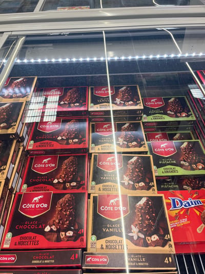 A refrigerated display case in a grocery store in Saint-Brice-Courcelles, France, stocked with boxes of "Côte d'Or" brand ice cream bars. The boxes are arranged in a grid pattern, each containing four ice cream bars. The ice cream flavors are chocolate, vanilla, and pistachio. The boxes all have the same design, with a gold background, a red or green band, and a picture of an elephant holding a scoop of ice cream. The price is 4.00 euros per box. The display case has an overhead light shining down on the ice cream. The display case is lit from above by fluorescent lights. The store's fluorescent lights are casting a warm glow on the scene. There is a box of "Daim" caramel ice cream to the right of the Côte d'Or ice cream bars. The Daim ice cream is packaged in a red and yellow box, with a picture of a caramel ice cream bar.