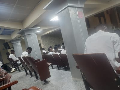 The image depicts an indoor scene, likely a hall or auditorium, in Kason, Nigeria. The environment features rows of dark brown chairs with metal legs, suggesting a seating area for an audience. Several individuals, predominantly young men wearing white shirts or garments, are seated in these chairs, facing towards a common direction, possibly a stage or presentation area that is not fully visible. The architecture includes large, gray, textured pillars and a suspended ceiling with a grid pattern. The lighting appears to be from overhead fixtures and natural light coming from windows or skylights, creating bright spots. The floor is a speckled gray tile. The overall atmosphere suggests a gathering or an event taking place in a formal setting.