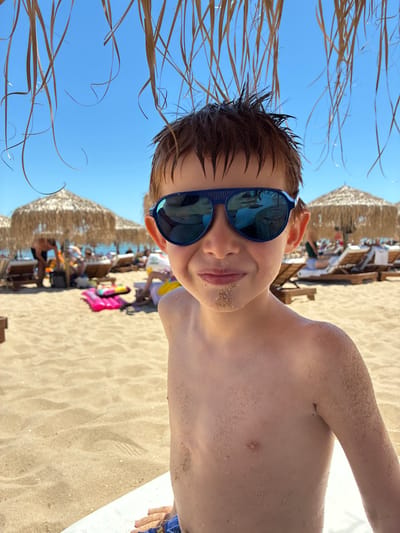 The image is a close-up, vertically oriented selfie of a young boy wearing blue sunglasses and covered in sand. He is on a beach, with the blue sky and dry grass visible above him, and the sandy beach with straw umbrellas and other beachgoers in the background. The boy appears to be happy and enjoying his time at the beach. The lighting suggests it is daytime, likely sunny and warm. The background shows a typical beach scene with people relaxing under umbrellas and the sand stretching out. There are no visible text elements in the image.