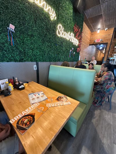 In Naples, Florida, at the Sunny House Lunch & Cafe, a brightly lit interior scene unfolds. The walls are adorned with a vibrant green artificial hedge, punctuated by glowing neon signs that read "Sunny" and "Start Happy." Red decorative berries add a festive touch. A table, set with menus and condiments, sits in the foreground. One menu features a "4th of July" theme, depicting various food items. A woman in a colorful floral dress is seated at a booth, conversing with other individuals. The atmosphere appears casual and inviting, typical of a cafe setting in the United States.