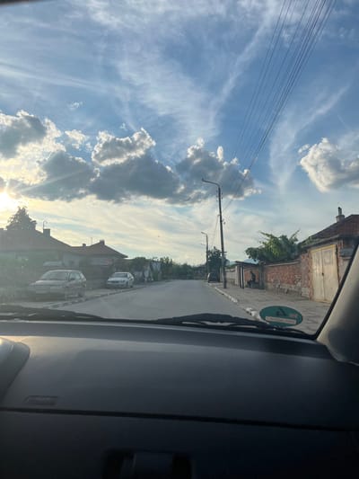 The image is taken from the interior of a vehicle, looking out through the windshield. The scene depicts a street in what appears to be a small town or village, with houses lining both sides of the road. The sky is a prominent feature, filled with dramatic clouds and streaks of sunlight, suggesting either sunrise or sunset. There are power lines running across the sky, supported by utility poles.
On the left side of the road, several parked cars are visible in front of houses. The houses themselves have red-tiled roofs and appear to be residential. On the right side of the road, there's a brick wall and a large garage door. A person is seated on a bench next to the brick wall.
The road is paved and seems to be in decent condition. The overall atmosphere is one of a quiet, possibly rural, setting bathed in the warm light of the sun.
Specifically, the image captures a street scene in Novo Zhelezare, Bulgaria. The architecture of the houses, the style of the parked vehicles, and the general ambiance are consistent with a location in rural Bulgaria. The presence of a person sitting on a bench along the sidewalk further grounds the scene in a specific, observable reality within the mentioned location.