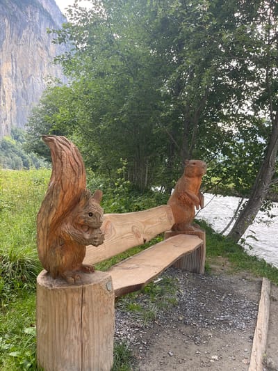 The image depicts a wooden bench with two carved wooden animal figures, a squirrel and a marmot, positioned on either side of the bench. The squirrel is on the left, carved from a tree stump, and the marmot is on the right, also carved from a tree stump. Both figures are intricately detailed, showcasing their fur texture and features. The bench itself is made of rough-hewn wood, with a natural, rustic appearance.
The scene is set in an outdoor environment, characterized by lush green vegetation, trees, and a flowing river in the background. A towering, steep cliff face dominates the left side of the image, likely a scenic backdrop. The ground around the bench is a mix of soil and gravel. The lighting suggests it is daytime, with sunlight filtering through the leaves of the trees.
The image was captured in Wengen, Switzerland. Specifically, this bench is located near the Staubbach Falls in the Lauterbrunnen Valley, a picturesque area known for its dramatic cliffs and waterfalls. The presence of the carved animals adds a charming, whimsical touch to the natural beauty of the Swiss Alps. There are no people or animals present in the image, and no discernible actions or interactions are taking place.