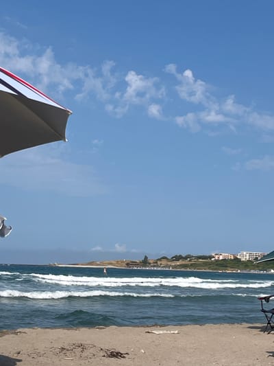 This media file depicts a scenic view of a beach in Ahtopol, Bulgaria. The scene is dominated by a bright blue sky with scattered white clouds, suggesting fair weather and likely daytime. The foreground features a portion of a beach umbrella, its dark fabric and red and white striped trim partially visible. To the right, the sandy beach is visible, with some dried seaweed or debris scattered on it. Beyond the beach, the turquoise waters of the sea are characterized by white-capped waves rolling towards the shore. In the distance, along the coastline, there's a strip of green vegetation and a cluster of buildings, possibly indicating a town or resort area. The overall impression is one of a peaceful and sunny day at the beach.