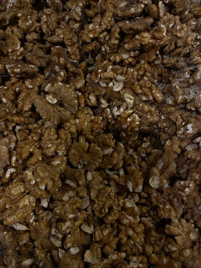 A close-up, top-down view of a large pile of shelled walnuts fills the frame. The walnuts are a rich brown color with lighter, irregular white patches on their surfaces. They are densely packed, with many pieces overlapping and some broken. The texture is rough and varied, with the characteristic convoluted shapes of walnut halves visible. There are no people, animals, or other objects present, and the background is entirely composed of more walnuts. The image is well-lit, highlighting the details and textures of the nuts. This image does not depict any location within the city of Perm, Russia.