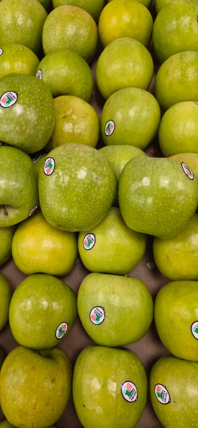 This is a close-up, eye-level, vertical shot of a large pile of green apples. The apples are densely packed together, filling the entire frame. Most of the apples are a vibrant green, with some showing yellow undertones. Several apples have small, circular stickers on them that appear to be branding labels. The background is a muted brown, suggesting a cardboard box or similar surface. The lighting is even, illuminating the textured surfaces of the apples. There are no people, animals, or discernable environmental features beyond the apples and their container. This image was likely taken in Navalcarnero, Spain, in a setting where fresh produce is stored or sold, such as a market, grocery store, or warehouse.