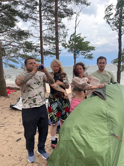 In Luzhki, Russia, on a sandy beach with a backdrop of the sea and overcast sky, four individuals and a dog are gathered around a partially set up green tent. The man on the left, wearing a floral short-sleeved shirt and black pants, is drinking from a blue bottle. To his right, a woman with blonde hair, in a black top and a floral skirt, holds a small brown dog. Next to her, a woman in a light pink sheer blouse over a green bikini top and pink shorts is in motion, possibly adjusting the tent, with her hair blowing in the wind. The fourth individual, a man in a green t-shirt and dark pants, stands to the right of the women, looking towards the viewer with a concerned expression. The scene suggests a casual outdoor gathering or camping trip.