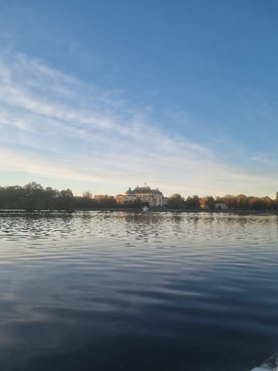 The image depicts Drottningholm Palace, located in Ekerö kommun, Sweden, viewed across a body of calm, rippled water, likely Lake Mälaren. The foreground consists of dark, undulating water exhibiting subtle reflections. In the midground, Drottningholm Palace, a large, light-colored, multi-story building with a complex dark roofline, stands prominently on a tree-lined shore. The palace's facade is illuminated by warm, low-angle sunlight. A small, white recreational boat is moored near the palace's shoreline on the left. Scattered deciduous trees and hints of other structures flank the palace. The background features a clear blue sky, lighter near the horizon and darker overhead, with sparse, elongated white cloud formations.