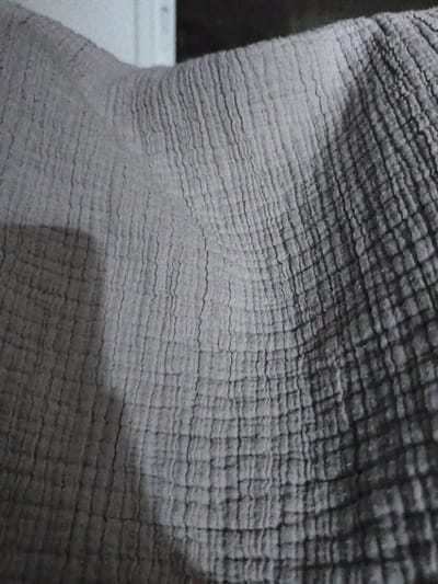 The image is a close-up, overhead shot of a grey, textured fabric. The fabric appears to be a gauze-like material with a crinkled or puckered surface, creating a pattern of raised ridges and valleys. The lighting casts shadows that highlight the texture and depth of the fabric. There is a shadow in the lower left quadrant of the image, suggesting a light source from the upper right. The background is out of focus and appears to be a white or light-colored object on the left and a dark, indistinct area on the right. No specific location in the city of Lod, Israel, can be identified from this image.