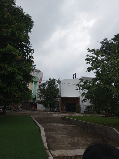 An outdoor scene in Cianjur, Indonesia, is depicted under an overcast sky. In the immediate foreground, the top of a person's dark-haired head is visible in the lower right. A concrete curb separates a strip of artificial green turf on the left from a worn, dark-colored path composed of dirt and concrete, which extends into the midground.
Lush green trees with dense foliage flank the path on both sides. In the midground, a group of indistinct individuals appears to be seated on the ground or low structures to the left, near some colorful objects.
Two distinct buildings dominate the background. On the left, a multi-story structure exhibits a unique architectural design with curved, pastel-colored sections (light green, yellow, white) and a rounded top section featuring a railing. To the right, a modern, white building with a curved facade is present. Its recessed entrance is dark, possibly composed of glass, beneath a brown-textured overhang, with a single exposed light bulb hanging above. On the flat roof of this white building, two dark, upright human-like figures are standing, facing away from the viewer.