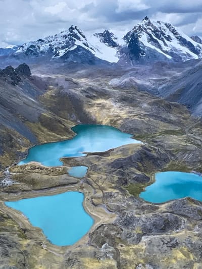 The image displays a breathtaking aerial view of a rugged, high-altitude mountainous landscape. Snow-capped peaks dominate the left side of the frame, their jagged, rocky slopes partially covered in white snow and ice, contrasting with the dark gray and brown rock. A soft, overcast sky is visible behind these peaks.

To the right, several vibrant turquoise-blue lakes are nestled within a desolate, rocky terrain. These glacial lakes vary in size and shape, with some interconnected by narrow channels of water, suggesting a flow between them. The striking blue color of the water is characteristic of glacial melt, rich in rock flour. The surrounding land is barren, composed of dark gray, brown, and olive-toned rocks, with sparse vegetation, indicative of an environment above the tree line. The texture of the terrain is rough and eroded, showcasing millennia of geological activity.

The scene depicts a natural, untouched wilderness. There are no people, structures, or signs of human activity visible. The lighting is diffused, suggesting either an overcast day or indirect sunlight, possibly late morning or early afternoon, highlighting the colors without harsh shadows. No text is visible in the image.

While a location context of Otopeni, Romania was provided, the visual evidence strongly suggests a high-altitude mountain range such as the Andes or Himalayas, given the snow-capped peaks, glacial lakes, and barren terrain, which are not characteristic of the region around Otopeni, Romania.