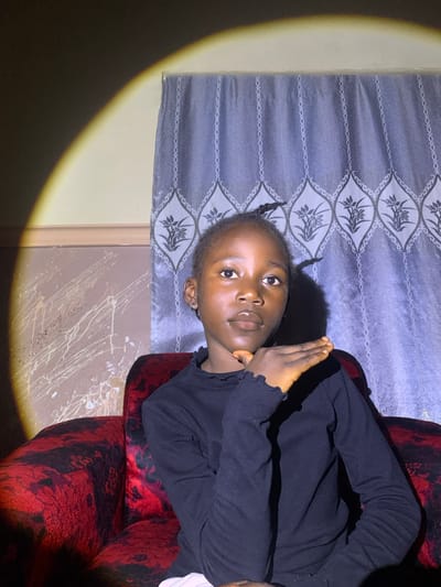 A young girl with braided hair is seated on a red armchair in what appears to be a room. She is wearing a black long-sleeved shirt. Her right hand is raised to her chin, and she is looking directly at the camera. A circular light is illuminating the scene from the left, casting a shadow on the wall behind her. The wall has a patterned wallpaper, and there is a grey curtain with a floral design behind the girl. The armchair is upholstered in a dark red fabric with a black floral pattern. The location is identified as Barakin Sabawa, Nigeria.