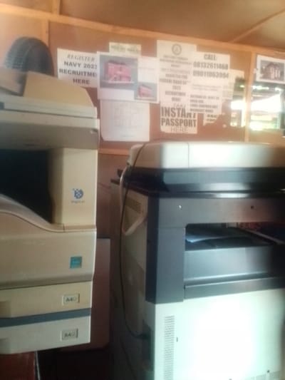 The image shows two photocopiers side by side in a small office located in Auchi, Nigeria at the coordinates latitude: 7.062, longitude: 6.273. The photocopier on the left is cream-colored with "A4" in blue lettering on two of its panels. The photocopier on the right is white and black, with the top being white, the middle being black and the bottom being white. The bottom of the right photocopier has a series of vertical lines on its front panel. There is a black cord running from the right photocopier to the left photocopier. The background is a wooden wall with a corkboard covered in posters. The posters advertise services for things like "instant passport," "navy recruitment," and "federal road safety recruitment." The top of the corkboard is made of wood. The bottom of the corkboard is covered with a piece of what looks like brown paper or fabric. There is a small window on the far right.
