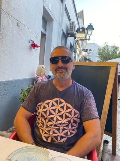 A medium shot captures a man with short, graying hair and a beard, wearing sunglasses, a dark t-shirt with a geometric pattern, and a bracelet, seated at a table outdoors in Olhão, Portugal. He is looking directly at the camera. Behind him, a woman with blonde hair is visible. The background shows a white building with a gray lower section, an air conditioning unit, a sign that reads "DIAMOND RESTAURANTE SNACK-BAR", a lamppost, and a blackboard on an easel. Plants are growing along the wall. The lighting suggests it is daytime, possibly late afternoon or early evening.