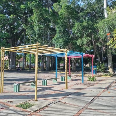 The image shows a park in Duaca, Venezuela, with a playground structure and surrounding trees. The playground structure is made of yellow, blue, and pink metal bars and has green benches underneath. The ground is paved with concrete and red bricks, and there are fallen leaves and grass growing in some areas. In the background, there are large trees, some with tall, slender trunks. A few people are sitting on a bench in the distance, and a broom is lying on the ground. The sky is clear and blue.