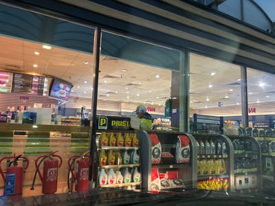 The image depicts a view through a glass storefront of a gas station convenience store. The store is well-lit with fluorescent ceiling lights and features shelves stocked with various products. In the foreground, three red fire extinguishers are visible, two of which have Cyrillic labels. Behind the fire extinguishers, shelves display an assortment of automotive fluids, including motor oil and other liquids in yellow, gray, and clear plastic bottles. There are also bags of charcoal visible on lower shelves.
Inside the store, a person wearing a high-visibility yellow jacket and a cap is standing at a counter, looking at something. The store appears to be a "VIVA Cafe" brand, with signage visible. A menu board displaying food and drink items is positioned on the left side of the store. The overall environment suggests a typical roadside convenience store located in Kalugerovo, Bulgaria, likely attached to a gas station, given the products displayed. The time of day appears to be evening or night, as evidenced by the interior lighting and the reflection on the glass.