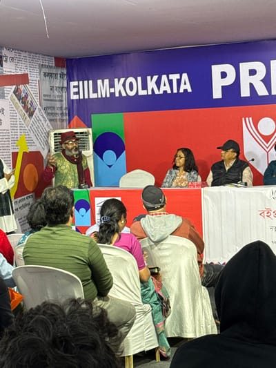 The image depicts an indoor event, likely a panel discussion or seminar, taking place in Bidhannagar, India. The backdrop features a large blue banner with "EIILM-KOLKATA PR" in white text, positioned above a red section that includes a white stylized logo resembling a person with raised arms. To the left of the stage, a wall is covered with Bengali newspaper clippings and abstract yellow and orange shapes; visible Bengali text on these papers includes "কলকাতা" (Kolkata) and "বইমেলা" (Book Fair), indicating the event is part of a book fair in the Kolkata metropolitan area, specifically Bidhannagar.

At a white-draped table on a raised platform, a male speaker stands on the left, wearing a red fedora-style hat, glasses, a dark beard, a gold-patterned green jacket, and a maroon scarf. He is actively gesturing with his right hand while speaking into a microphone positioned on the table. To his right, a female participant with glasses and a patterned top is seated behind the table, alongside a male participant wearing a black baseball cap, glasses, and a dark sleeveless vest over a light shirt. A fourth individual in a blue shirt is partially visible to the far right. A white cooling unit is situated behind the standing speaker.

The foreground and midground show an audience seated in white plastic chairs, viewed primarily from behind. Visible audience members include a person in a green striped long-sleeved shirt, a person with dark hair tied in a ponytail wearing a pink top, and an individual in a brown hoodie with a striped hood and a black beanie. The audience is oriented towards the platform, attending the event.