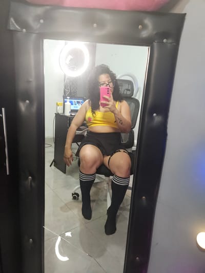 A woman is taking a mirror selfie in a room. She is sitting on a black office chair in front of a desk. She is wearing a yellow crop top, a black mini-skirt, and black knee-high socks with white stripes. She has curly dark hair and is wearing glasses. She is holding a pink smartphone. In the background, there is a ring light, a laptop, and bottles on the desk. The room appears to be a home office or gaming setup, with a tiled floor. The mirror she is using to take the selfie has a black, padded frame.