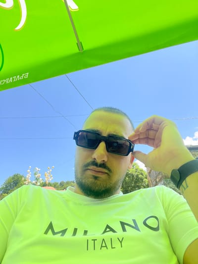A close-up, low-angle portrait shot captures an adult male outdoors, identified as being in Касапите, Bulgaria. The man looks directly into the camera. He has short, dark hair, a beard, and a mustache. His skin exhibits a yellowish hue, consistent with illumination reflected from the large, bright green umbrella positioned directly above him, which occupies the top third of the frame. The umbrella's underside displays reversed white and orange/yellow Cyrillic text, reading "ЕРУЛЧВСН" (implying "НСВЛЧУРЕ").

The man wears dark, thick-framed rectangular sunglasses with a small, gold-colored detail on the outer edge of the right temple. His right hand (viewer's left) is raised, with fingers gently touching the right temple of his sunglasses, as if adjusting them. On his left wrist (viewer's right), he wears a dark, mesh-patterned wristband or watch, and a faint tattoo is visible on his inner forearm below the wristband. He is dressed in a neon green/yellow crew-neck t-shirt featuring the words "MILANO ITALY" printed in dark grey/black; "MILANO" is in a larger font above "ITALY."

The background reveals a clear, light blue sky traversed by several thin, dark horizontal lines, likely utility wires. Below the horizon line formed by the umbrella, green foliage from trees and bushes is visible. In the mid-ground, light-colored, decorative botanical elements resembling dried flowers or seed pods on slender stems are present. A light-colored stone or concrete structure is partially visible on the far right edge of the frame. The overall scene suggests a sunny day.