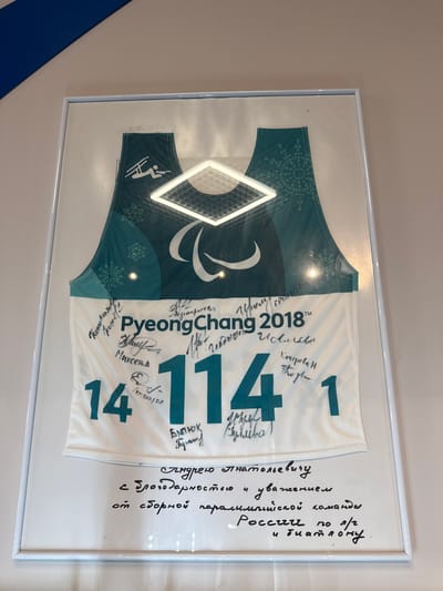 The image shows a framed jersey, likely a biathlon or cross-country skiing jersey, on display in what appears to be an indoor environment. The jersey is signed by multiple individuals and features the "PyeongChang 2018" Paralympics logo. The jersey number is "141". Written in Russian on the right side of the frame is a message addressed to Andrei Anatolyevich with gratitude and respect from the Russian Paralympic team for biathlon and cross-country skiing. Based on the information, this image is highly probable to be taken in the city of Peresvet, Russia.