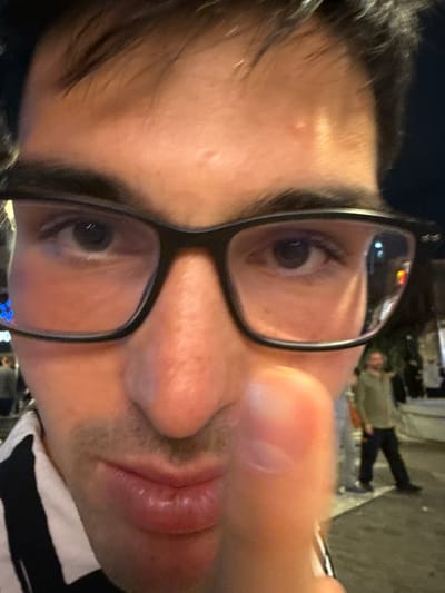 A close-up selfie of a man with glasses. The background is a street scene, likely in Thessaloniki, Greece, indicated by the reflections in the glasses appearing to show buildings and lights. Other people are seen in the background. The man has dark hair and light skin. He is wearing a black and white striped shirt. His expression is neutral. The photo appears to have been taken at night.