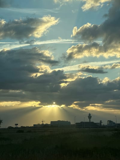 The image depicts a dramatic sunset over a landscape that includes an airport control tower and other airport buildings, consistent with the location of Schiphol Airport near Hoofddorp, Netherlands. The sky is filled with a mix of dark, ominous clouds and bright, sunlit clouds, with visible sunbeams piercing through the darker formations. The sun itself is partially obscured by clouds, casting a warm, golden light. In the foreground, there is a grassy field with some indistinct vegetation. The middle ground features a line of airport structures, including a prominent control tower, illuminated buildings, and what appears to be a construction crane. The horizon is marked by the silhouette of these buildings and some distant trees. The overall impression is one of a striking natural phenomenon occurring over a man-made infrastructure. There are no people visible in the image.