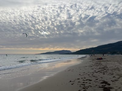 This image captures a beach scene in Tarifa, Spain, characterized by a dramatic sky filled with textured, fragmented clouds illuminated by diffused sunlight. The foreground is a sandy beach with scattered dark seaweed. The ocean stretches towards the horizon, with small waves breaking along the shore. Several individuals are visible on the beach and in the water. One person is kitesurfing in the distance, and another is on a paddleboard closer to shore. On the beach, several people are walking or standing, and one person is lying down on the sand, wearing an orange life vest. In the background, a hilly coastline with vegetation forms the horizon, and more kites are visible in the sea. The overall atmosphere is serene, with the soft light and the presence of water sports suggesting a typical day at this popular windsurfing and kitesurfing destination.