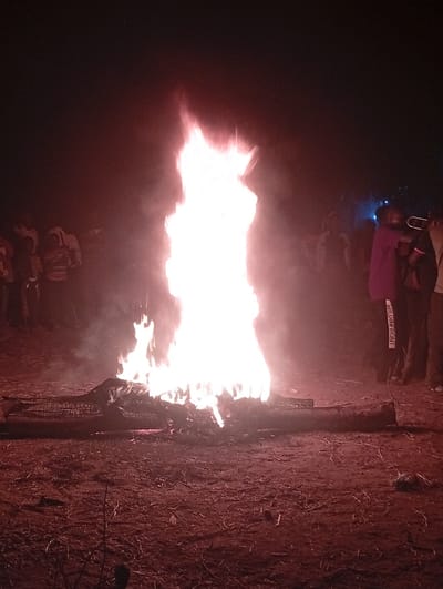 A large bonfire illuminates a dark scene at night, casting an orange-red glow on the surrounding ground. People stand around the fire, their figures silhouetted against the bright flames. Logs are visible beneath the fire. Some individuals are wearing hoodies. The scene suggests a gathering or event centered around the fire.