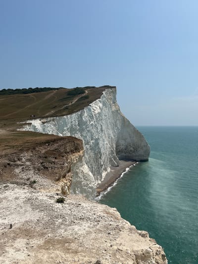 The image depicts a coastal landscape with dramatic white chalk cliffs meeting the turquoise sea. The cliffs are steep and sheer, with visible strata of chalk rock. A narrow strip of pebbly beach is visible at the base of the cliffs. The water is clear and appears to have varying shades of green and blue. On top of the cliffs, there is a grassy, undulating terrain with several dirt paths winding across it. A few small figures of people can be seen walking along these paths, and in the distance, a faint white sailboat is visible on the horizon. The sky is clear and a pale blue, with a few distant birds visible as tiny specks. The foreground of the image shows a rocky, eroded cliff edge with sparse vegetation. This scene is characteristic of the Seven Sisters cliffs near Seaford in East Sussex, United Kingdom.