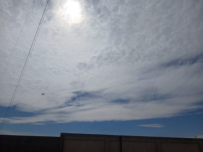 A slightly overcast sky with thin, wispy clouds is visible above a muted brown wall. The sun, partially obscured by clouds, shines brightly in the upper left quadrant. Two thin, dark lines, possibly wires, extend diagonally across the sky from the upper left towards the center. The wall appears to be constructed of concrete or plaster and forms the lower boundary of the image. The scene suggests an outdoor environment in a city.