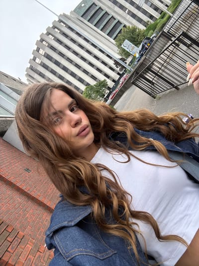 A young woman with long, wavy, light brown hair is taking a selfie outdoors. She is wearing a white t-shirt and a blue denim jacket. Her left hand is visible in the upper right corner of the frame, with long, white-tipped fingernails. The background features a modern building with a grid-like facade, a black metal fence, a street with parked cars and a street sign, and a brick wall in the foreground. The scene appears to be in an urban environment, likely a city. The specific location identified is the area near the Lighthouse Centre for the Arts in Poole, United Kingdom, as indicated by the street sign which points to "The Lighthouse".