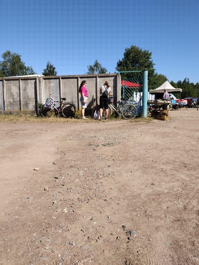 The image depicts an outdoor scene, likely a flea market or an informal gathering, under a clear blue sky. In the foreground, a dirt and gravel path fills the lower half of the frame. Behind this path, a concrete wall stretches horizontally across the image.
Along the wall, several bicycles are visible. One bicycle, on the left, has a dartboard attached to its spokes, with colorful points. Two women are standing near bicycles. The woman on the left wears a bright pink t-shirt and light-colored shorts. The woman on the right wears a striped t-shirt and dark shorts, and has a backpack. Further to the right, a green metal gate stands open, revealing a glimpse of a red canopy behind it.
Beyond the gate and the concrete wall, a market stall is set up under a white tent. A person, appearing to be a vendor, is behind the stall, interacting with a customer. Various items are displayed on tables at the stall, suggesting goods for sale. Several vehicles, including vans, are parked in the background, and a dense line of trees forms the horizon. The overall environment suggests a sunny day at a public gathering space. The scene is located in the city of Irshansk, Ukraine.