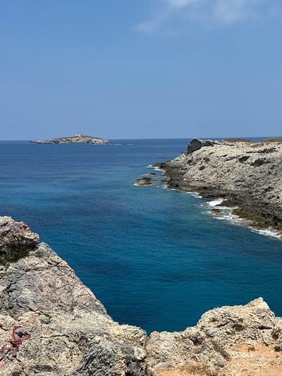The image captures a coastal scene with rugged, rocky terrain meeting the deep blue sea. In the foreground, large, weathered rocks with visible cracks and some red spray paint mark the edge of a cliff. The rocks are a pale, sandy color with gray and pinkish hues. Beyond the immediate foreground, the sea stretches out, its color transitioning from a vibrant turquoise near the shore to a deeper indigo further out.
A prominent feature in the middle ground is a rocky coastline that curves into the sea, with waves gently breaking against its base. Further in the distance, a small, elongated island with rocky outcrops and sparse vegetation sits on the horizon under a clear, pale blue sky. The overall impression is one of a remote and natural coastal landscape. The location is identified as being in the vicinity of Rizokarpaso, Northern Cyprus. No people or specific structures are visible in the image.