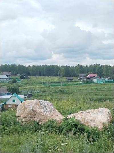 The image shows a rural landscape with a cloudy sky. In the foreground, two large, light-colored boulders sit on a grassy slope. Behind the boulders, a village with several houses and outbuildings is visible. The houses are a mix of styles, with some having green roofs and others painted in various colors. A dense forest borders the village in the background. The overall impression is one of a peaceful, rural setting in the village of Idrisovo, Russia.