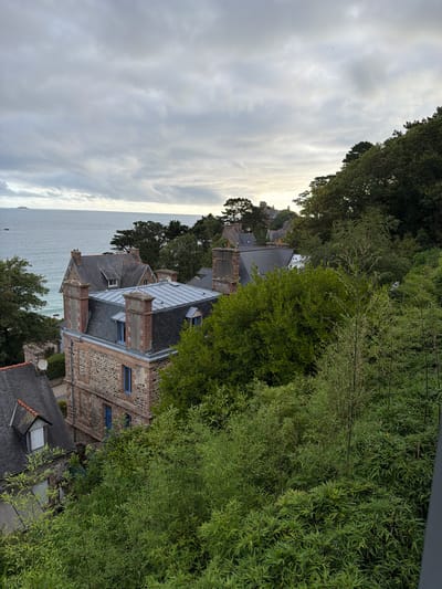 The image is an elevated, eye-level exterior shot of the coastal town of La Clarté, France, under a cloudy sky. The scene features several stone and slate-roofed buildings with red brick accents, nestled amongst lush green trees and vegetation. The ocean is visible in the background, with a small island in the distance. The architecture suggests a historical European coastal village. There are no people or animals present.