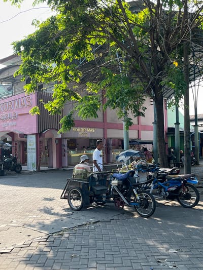 This image depicts a street scene in Juwet Kenongo, Indonesia. The foreground is dominated by a paved street with visible cracks and uneven sections. A three-wheeled vehicle, possibly a modified motorcycle or a small cart, is positioned in the lower center, featuring a blue fabric cover and a person wearing a light-colored hat and a green garment seated on it. Next to it, a blue motorcycle is parked.
In the middle ground, two men are standing near the three-wheeled vehicle. One man, wearing a white shirt, appears to be interacting with the other man who is seated. Behind them, a pink building with "BOKOR MAS GOLD EST 1978" signage is visible. The building has a striped facade and several entrances. A vertical sign with text and graphics is also displayed on the building's exterior. Several motorcycles are parked in front of the building.
The background reveals more of the street and buildings, with some trees providing shade. The overall atmosphere suggests a commercial area, likely a market or a street with shops. The lighting indicates it is daytime, with sunlight casting shadows on the pavement.