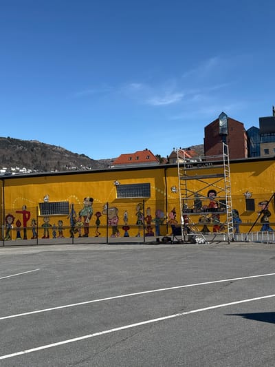 The image, correctly oriented, depicts a sunny urban scene in Bergen, Norway, dominated by a vibrant yellow building. The long building features a colorful, cartoon-style mural along its lower facade, portraying numerous whimsical figures, possibly children or stylized characters, in a lively, continuous arrangement. Small, illustrative elements like birds and dotted lines are interspersed within the mural. Several rectangular windows, appearing to be glass blocks, are also visible on the yellow wall.
In the foreground, a grey asphalt surface, likely a parking lot or road, extends from left to right, marked with crisp white lines. A metal scaffolding structure is erected against the upper part of the yellow building, partially obscuring some windows and sections of the mural, suggesting ongoing construction or maintenance, though no workers are present.
Beyond the main yellow building, several other structures are visible, including a modern reddish-brown brick building with prominent glass features, and further back, traditional houses with red-tiled roofs. A distinctive mountain, characteristic of the Bergen landscape, rises in the background, sparsely covered with vegetation.
The scene is set on a bright, clear day, likely midday or early afternoon, evidenced by the strong sunlight and defined shadows on the asphalt. The sky is a deep blue with only a few faint, wispy clouds. The mood conveyed is cheerful and bright, primarily due to the mural's playful nature. No discernible text is visible in the image.