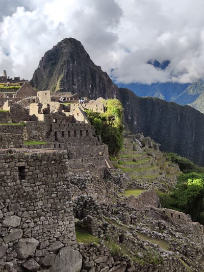 Machu Picchu is shown in a landscape under partly cloudy skies. The Incan ruins consist of stone structures and walls on multiple levels, built into the surrounding mountainous terrain. A prominent peak looms in the background, its upper reaches shrouded in clouds. Terraced slopes are visible. The structures are illuminated by natural light, casting shadows.