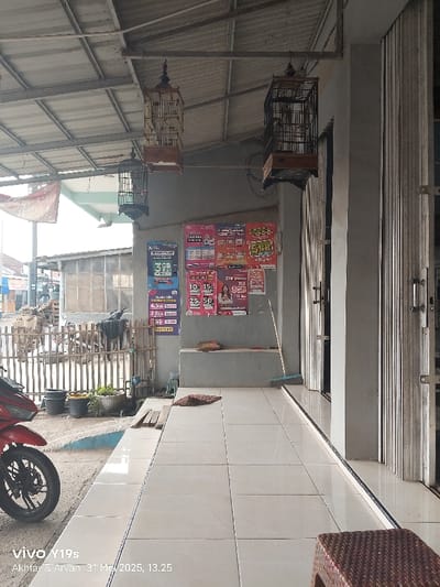 The image shows the exterior of a building located in Kecamatan Anjatan, Indonesia, at the coordinates provided. The scene is captured from a low angle, showcasing the tiled entrance area with a small, brown wicker stool. Three birdcages are hanging from the roof overhang, two of them visible in the frame. The building's walls are a dull grey and feature a series of promotional posters in Indonesian. A wooden fence separates the building from the exterior area, which is partially paved and features a red motorcycle parked in the foreground. There is a small potted plant next to the motorcycle and a few other plants are visible in the background. The overall image is of a quiet, ordinary scene in an urban area.