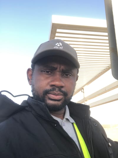 The image is a close-up, selfie-style outdoor portrait of an adult male located in Hanak, Saudi Arabia. The man has a dark complexion, a dark beard and mustache, and is looking directly at the camera. He is wearing a dark grey baseball cap with white Arabic script ("أمرك") and a small white emblem on the front. He is dressed in a black jacket, partially unzipped, revealing a light grey collared polo shirt and a neon yellow reflective strip underneath on his left side. The background consists of a light-colored, possibly metal or concrete, overhead structure with multiple horizontal slats and a vertical support column, set against a clear, bright blue sky, indicating a daytime scene.