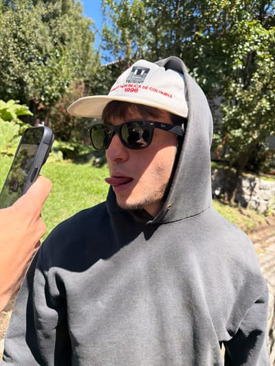 A person wearing a gray hoodie and a cap with "Tecnint Republica de Colombia 1996" printed on it. They are also wearing sunglasses. Their tongue is sticking out. They are holding a smartphone in their hand, possibly taking a selfie. The background includes green trees and a hint of blue sky, suggesting an outdoor setting. The photo appears to be taken at an angle.