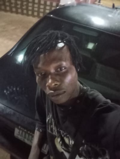 A nighttime, low-light photograph depicts a young adult male in Onicha, Nigeria. The individual, of dark complexion, has dark hair styled in dreadlocks, with a white band or object across his forehead. He wears a dark long-sleeved shirt with a light-colored graphic on the chest. He is positioned in front of a dark-colored sedan, with the vehicle's front hood and windshield visible. A blurry, reversed sequence of characters, consistent with a license plate, is discernible on the lower front of the car. The background is out of focus, showing indistinct architectural elements and faint light sources, indicating an urban outdoor setting after dark.